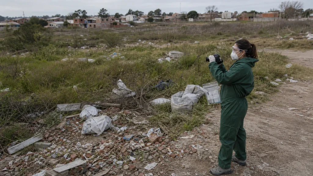 Trabajadora de salud ambiental documentando un microbasural periurbano con protección respiratoria.